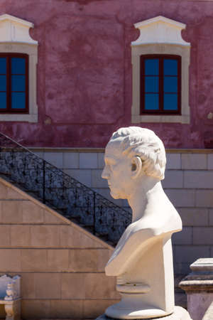 Estoi, Portugal - June 15, 2018 Bust from a profile, made of a white stone. Line of the staircase and typical bordeaux older facade of the Palace, with windows reflecting blue sky.のeditorial素材