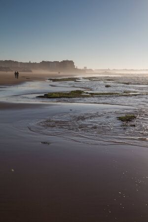 Sandy beach with small rocks in the late afternooon. Water of Atlantic ocean. Silhouette of the city in a misty background. Blue sky. Essaouira, Morocco.の写真素材
