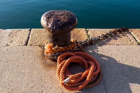 Concrete pathway with a rusted old bollard and an orange rope. Calm water of Mediterranean sea in...