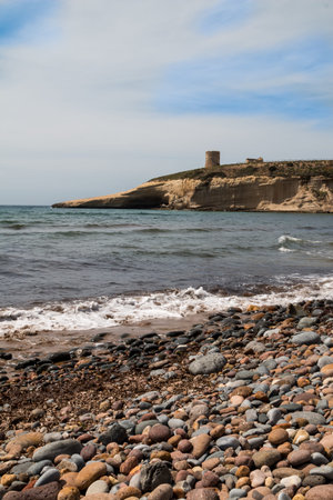 Beach in the bay, covered by sand and pebbles. White foam on the waves of the Mediterranean sea. Round of a mountain, creating the bay, with a historical defence military tower. West Sardinia, Italy.の写真素材