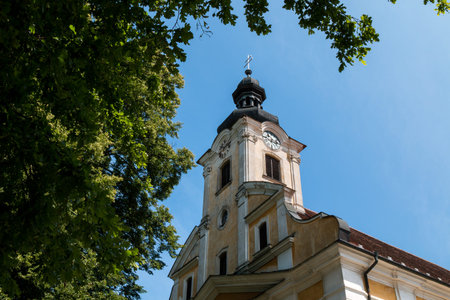 Weathered yellow facade with white details of the Saint Stephen (Svaty Stefan) church. Tower with clock. Tree with fresh leaves in the spring. Blue sky. Jablonica, Slovakia.の写真素材