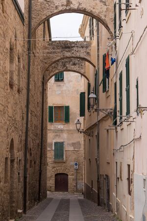 Old narrow street with arc in between of the houses. Windows with shutters. Hanging laundry. Downtown of Alghero, Sardinia, Italy.の写真素材