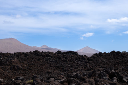 Lava soil of a brown to black color. Mountains in the background. Blue sky with white clouds in the early spring. Lanzarote, Las Palmas, Canary Islands, Spain.の写真素材