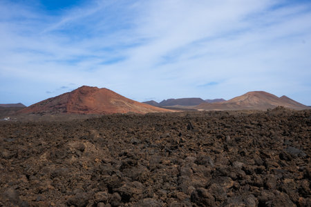 Lava soil of a brown to black color. Mountains in the background. Blue sky with white clouds in the early spring. Lanzarote, Las Palmas, Canary Islands, Spain.の写真素材