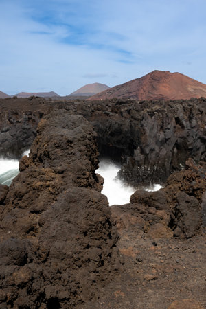 Dark brown to black color of the lava stones and rocks, created a cliff above the wild water of the Atlantic Ocean. Cloudy sky in the early spring. Mountains in the background. Los Hervideros, Lanzarote, Canarian Islands, Spain.の写真素材