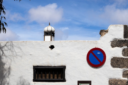 Old wall painted white with a small ventilation window and a No parking sign. Behind the wall top of the church tower. Blue sky with white clouds. Teguise, Lanzarote, Canary Islands, Spain.の写真素材