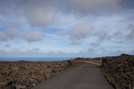 National Park of the volcanic formations. Colors of the land from red and brown to black. Just a road with limited traffic to preserve the heritage. Blue sky with white clouds. Timanfaya, Lanzarote, Canary Islands, Spain.の写真素材