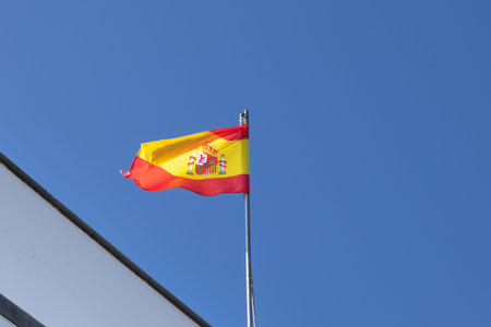 Triangle of a roof of a building and a pole with spanish flag, blowing in the wind. Bright blue sky....