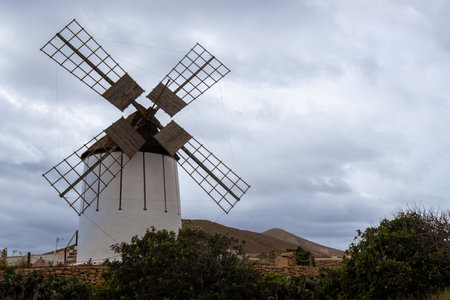 White facade of the windmill base. Located in the nature, between plants and bushes. Mountains in the background. Cloudy sky. Tiscamanita, Fuerteventura, Canary Islands, Spain.の写真素材