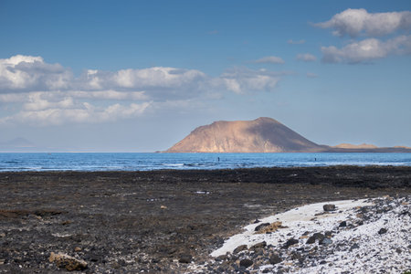 Dark lava soil on the beach and white pieces of coral. Calm Atlantic ocean. Volcanic mountain Lobos...