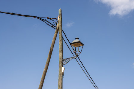 Old wooden pole with more cables and a traditional lantern - street light. Bright blue sky and light white clouds. Casillas del Angel, Fuerteventura, Canary Islands.の写真素材