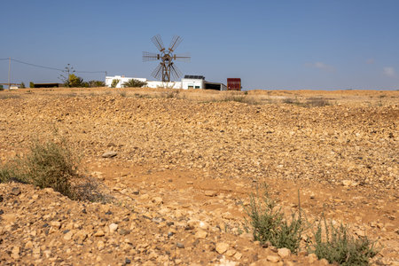 Sandy ground full of stones and rocks. White building of a farm with a windmill. Blue sky. Almacigo,...