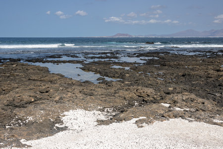 Dark rocks on the beach of Atlantic ocean. White areas filled with pieces of broken corals in the shape of popcorn. Blue sky with light white clouds. Playa del Bajo de la Burra and Playa el Mejilon, Fuerteventura.の写真素材