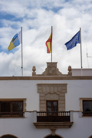 White building of a town hall. Traditional spanish architecture.Three  flags on the roof blowing in the wind: European Union, Spanish, Canary Islands. Puerto del Rosario, Fuerteventura, Spain.の写真素材