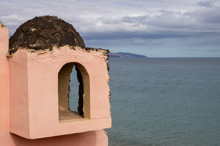 Orange building on a hill above the beach. Calm water of the Atlantic ocean. Mountains in the background. Cloudy sky in the winter. Costa Calma, Fuerteventura, Canary Islands, Spain.の写真素材