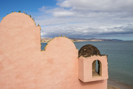 Orange building on a hill above the beach. Calm water of the Atlantic ocean. Mountains in the background. Cloudy sky in the winter. Costa Calma, Fuerteventura, Canary Islands, Spain.の写真素材