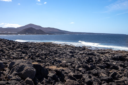 Detail of a textured dark brown to black volcanic stones on the coast of Atlantic ocean. Mountain in the background. Blue sky with some white clouds. Central Lanzarote, Canary Islands, Spain.の写真素材