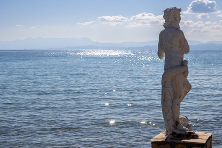 Calm and clean Ionian sea. Statue of Nereus in the harbor. Glittering water. Blue sky with light clouds. Mountains of Corfu in the background. Island Erikousa, Greece.の写真素材