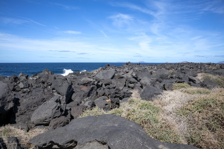 Dark rocks on some part of the Atlantic ocean shore. Calm blue water. Blue sky with light clouds. Lanzarote, Canary Islands, Spain.の写真素材