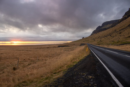 Black coastal road. Dry autumn grass around. Road passing by mountains on its right side. Cloudy...