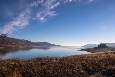 Sunny day with a blue sky and light clouds. Fjord surrounded by meadows and mountains with an autumn color of the vegetation. Eskifjordur, East Iceland.の写真素材