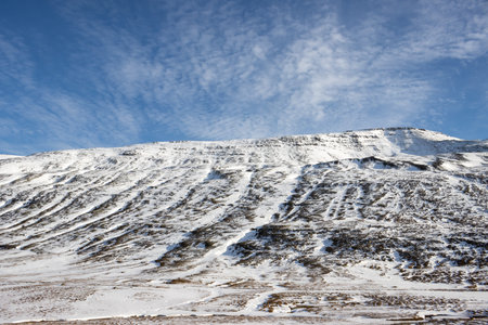 Rough dark volcanic rocks, covered with small layer of a snow, underlining how rough they are. Bright blue sky, light white clouds. Fjardabyggd, East Iceland.の写真素材