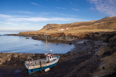 Small port in a fjord. Old abandoned rusted fishing boat beside. Water lined by mountains. Blue sky with white clouds. Bakkagerdi, Iceland.の写真素材