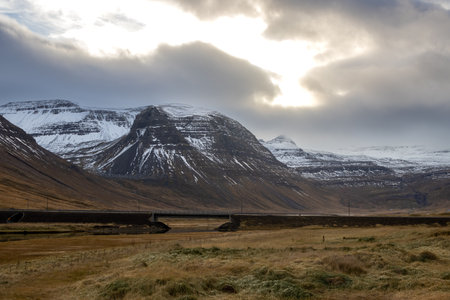 Majestic mountain with a touch of a snow in the autumn. Cloudy sky with a little sunshine. River in the valley. Bridge across the river. Area of Sydridalur, Westfjords, Iceland.の写真素材