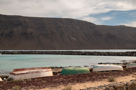 Dark brown cliffs on the north of Lanzarote. Shallow water of a beach. Boats on a beach. Blue sky with white clouds. Caleta del Sebo, La Graciosa, Canary Islands, Spain.の写真素材