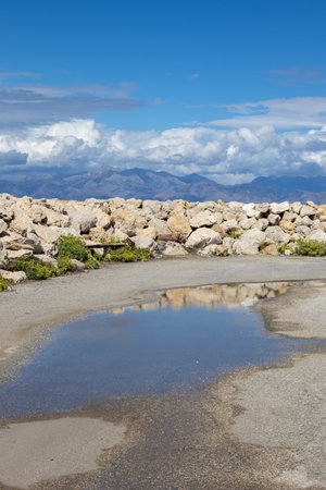 Large puddle after a rain. Pier with a stone breakwater wall. Albanian mountains in the background. Blue sky with white clouds. Astrakeri, Corfu, Greece.の写真素材