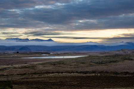 Autumn nature. Ground covered by grass, moss and lichen. Water of a lake. Mountains with a fog in the background. Sunset cloudy sky. Foraging / Hvalfjordur, Iceland.の写真素材