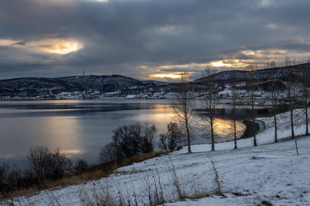 Curve of Kwaefjord, linded by landscape covered by snow in the time of sunset. Cloudy sky with little sunny spots, reflected on the water. Trees and bushes. Nupen, Norway.の写真素材