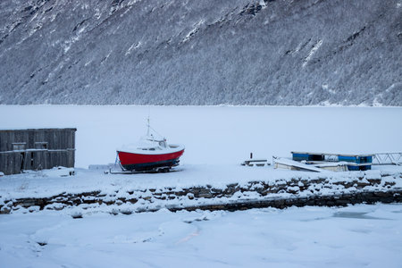 Red small boat on the coast of a fjord. Majestic mountain enlighted by sunlight and covered by snow in the background. Blue sky.の写真素材