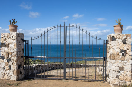 Metal ornate gate with stone pillars and pots with cactuses. Blue calm water of the Mediterranean sea.  Blue sky with white clouds in the spring. Portopalo, Sicily, Italy.の写真素材