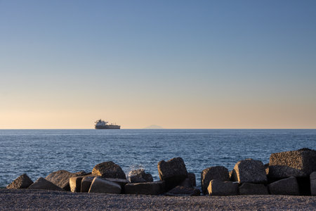Calm dark blue water ot the Tyrrhenian (Mediterranean) sea. Breakwaters on the beach. Big ship and Aeolian islands on the horizon. Blue sky with orange light in the early sunset. Caldera, Sicily, Italy.の写真素材