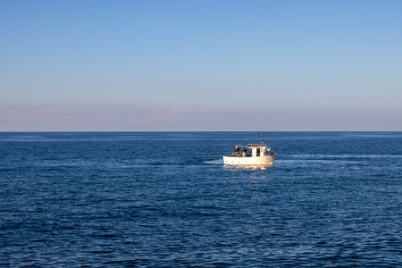 Calm dark blue water ot the Tyrrhenian (Mediterranean) sea. Small white boat. Blue sky with pink tones on the horizon with starting sunset. Caldera, Sicily, Italy.の写真素材