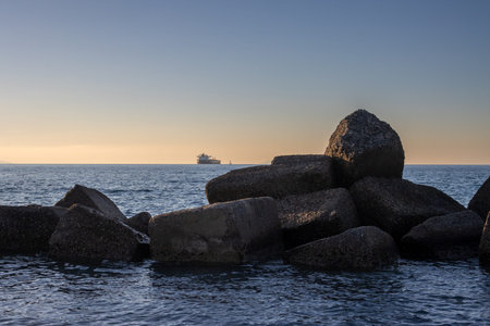 Beach with breakwaters. Calm blue water ot the Tyrrhenian (Mediterranean) sea. Ship on the horizon. Blue sky with orange tones during sunset. Spadafora, Sicily, Italy.の写真素材