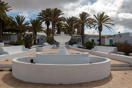 Park on a square with white built fountain and benches aroung. Palm trees and houses in the background. Blue sky with intense white clouds in the winter. Teguise, Lanzarote, Canary Islands, Spain.の写真素材