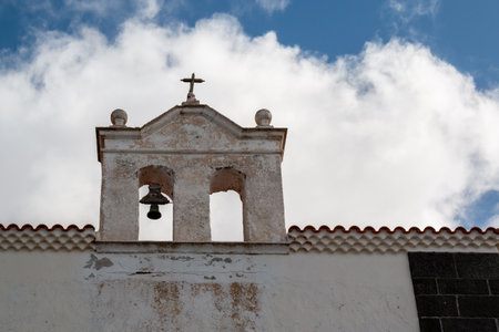 Detail of a belfry of an old church of the Hermitage of  Veracruz.Black and white facade. Belfry with one bell and a simple cross. Blue sky with white clouds. Teguise, Lanzarote, Canary Islands, Spainの写真素材