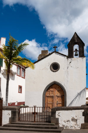 White facade of a chapel from 15.century. Belfry with one bell and a cross on the top. Blue sky with white clouds. Funchal, Madeira, Portugal.の写真素材