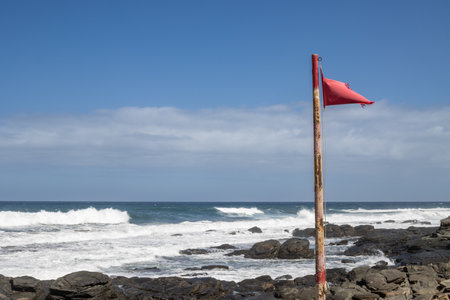 Red flag as a symbol of prohibited swimming on the rocky beach of Atlantic ocean with big waves. Blue sky with white clouds. Las Palmas, Gran Canaria, Canary Islands, Spain.の写真素材