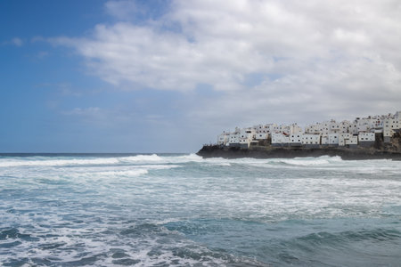Big waves of the Atlantic ocean. Town built on a archipelago rock. White houses. Cloudy sky. El Roque, Las Palmas, Gran Canaria, Canary Islands, Spain.の写真素材