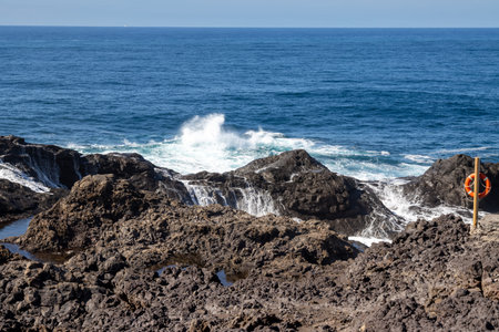 Dark volcanic coastal rocks. Calm Atlantic ocean with waves on the line of rocks. Blue sky. Galdar,...