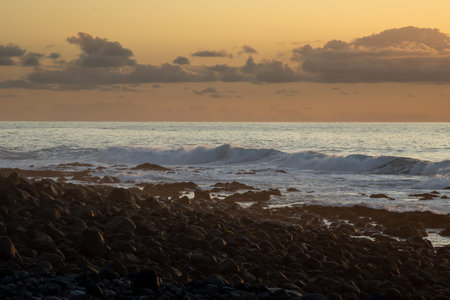 Volcanic pebbles on the beach of Atlantic ocean with waves. Orange sky with clouds during sunset....