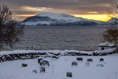 Tombstones at a cemetery, graves covered by snow. Stone fence. Norwegian sea and a mountain on the horizon. Colorful sky in an arctic sunrise, Harstad, Norway.の写真素材