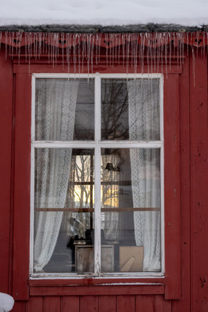 Traditional red wooden house. Window with white thin frame and a curtain, view on the opposite window. Icicles on the edge of the roof with snow. Harstad, Norway.の写真素材