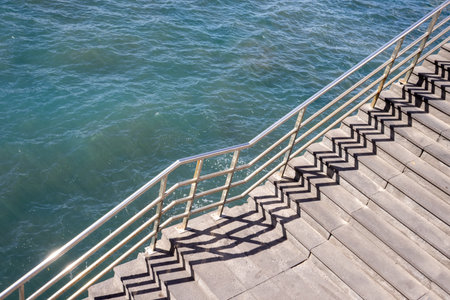 Diagonal view on a staircase with a railing, which is creating a shadow on the stairs. Green-blue water of Atlantic ocean in the background. Tenerife, Canary Islands, Spain.の写真素材