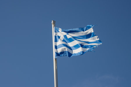 Flag on a flagpole outdoor, blowing in the summer wind. Bright blue sky in the background. Paxos, Greece.の写真素材