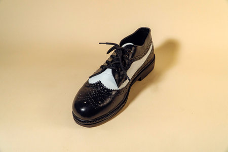 Close up of black and white brogue wingtip shoes with rubber outsole made of genuine cowhide. Studio shot of an elegant & shiny vintage two tone shoe product on a beige backgroundの写真素材