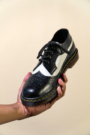 A man's hand holds a black and white brogue wingtip shoe with a rubber outsole made from genuine cowhide. Men's hands holding elegant and shiny two tone shoes on a cream backgroundの写真素材